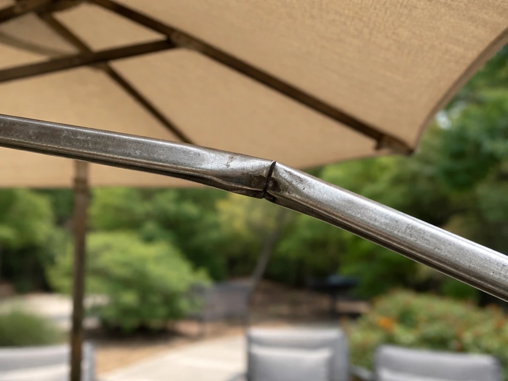 Close-up of a patio umbrella rib kinked mid-span, showing the exact failure point.
